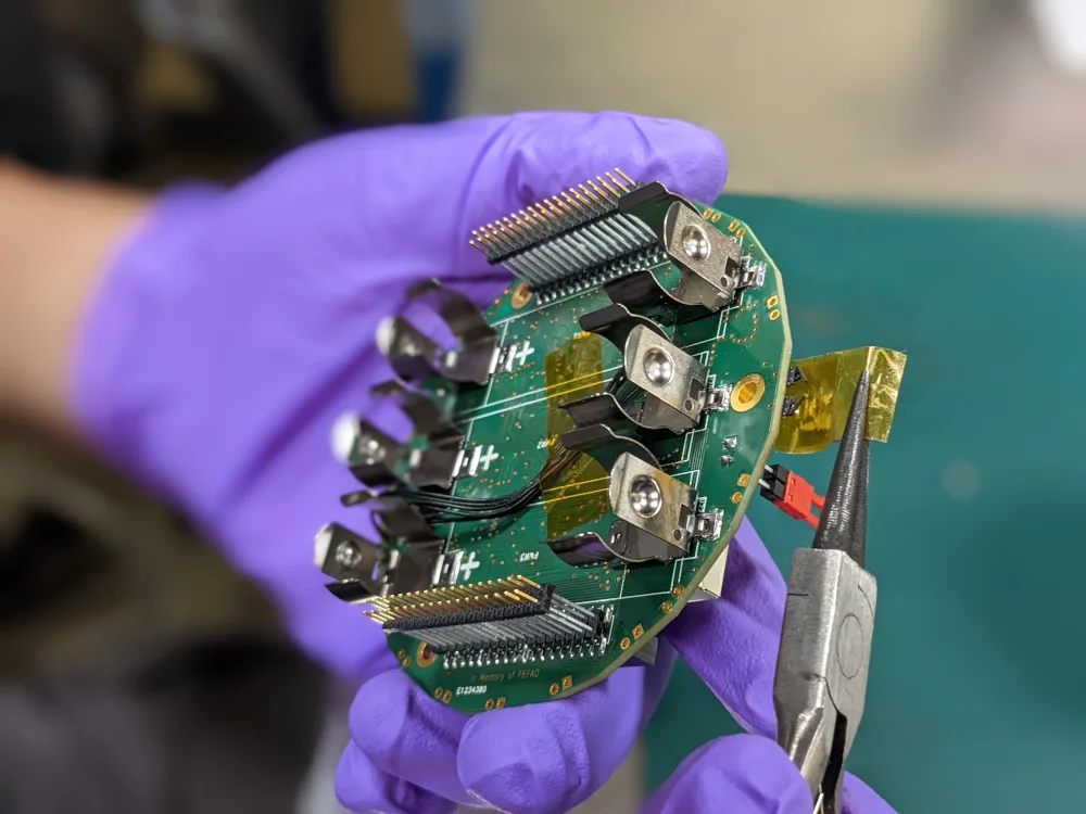 Lab member Aaron Gonzalez seated at a workbench, soldering and inspecting BobcatOrbiter's main avionics board.